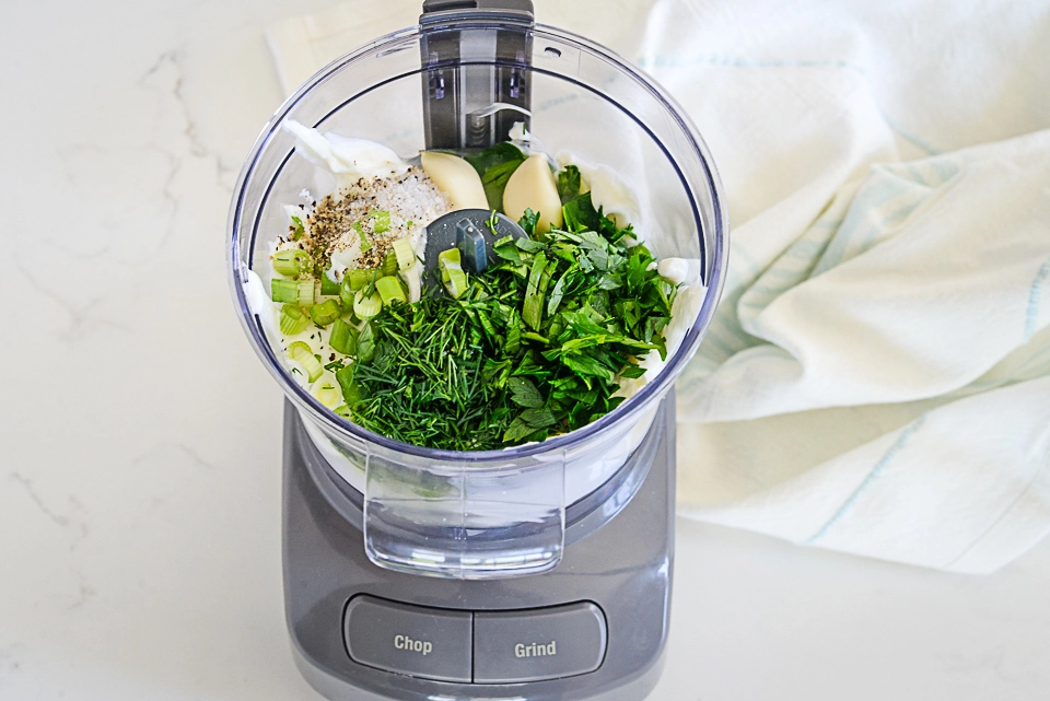 Inside view of a food processor with herbs, garlic, and seasoning being prepared for the wrap sauce.