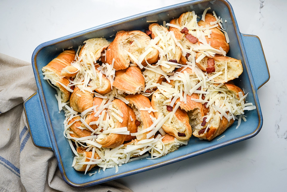 Shredded cheese layered over torn croissants and bacon bits in a blue casserole dish.