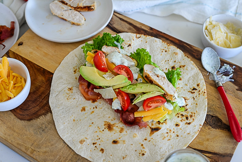 Grilled chicken wrap assembly with avocado, cheese, and sauce on a wooden board.